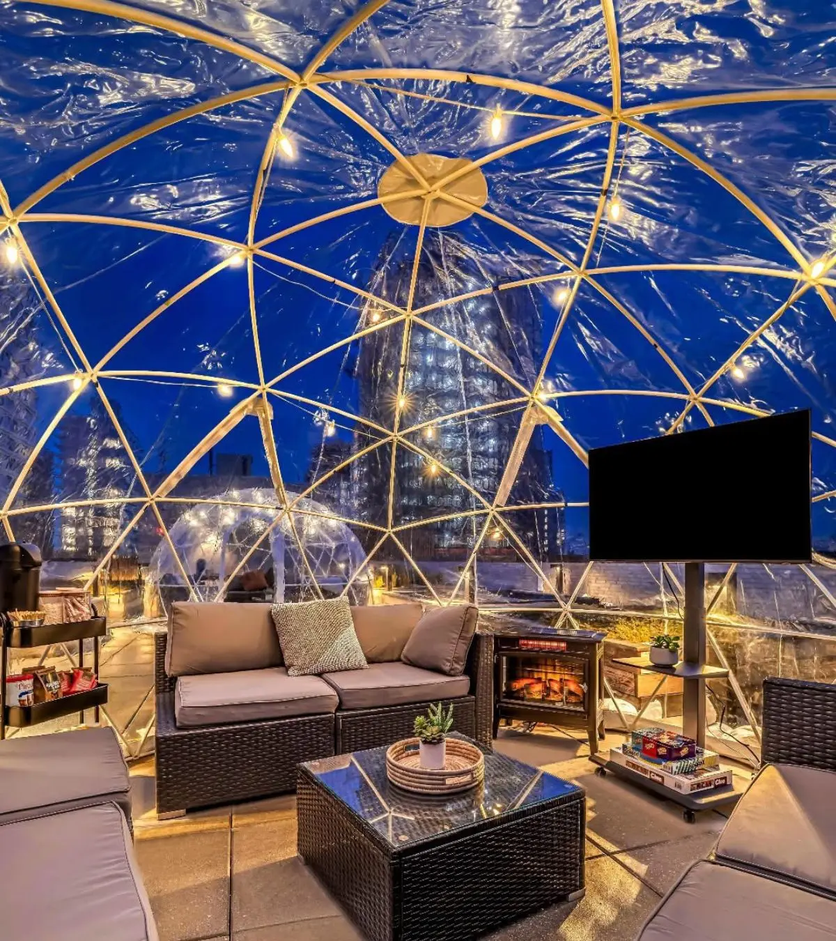 Furnished heated igloo interior on a New York City rooftop with lounge seating, lighting, and electric fireplace