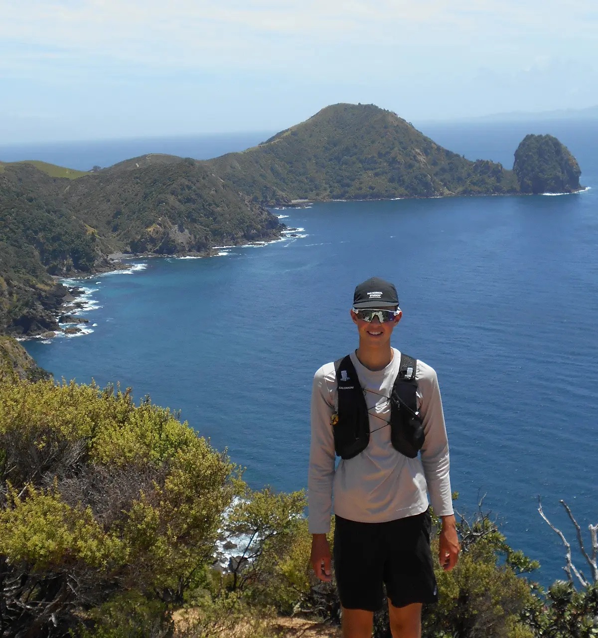 Junger Mann (Ferry Commentz) in Sportkleidung und Sonnenbrille vor einer felsigen Küstenlandschaft mit blauem Meer und bewachsenen Hügeln.