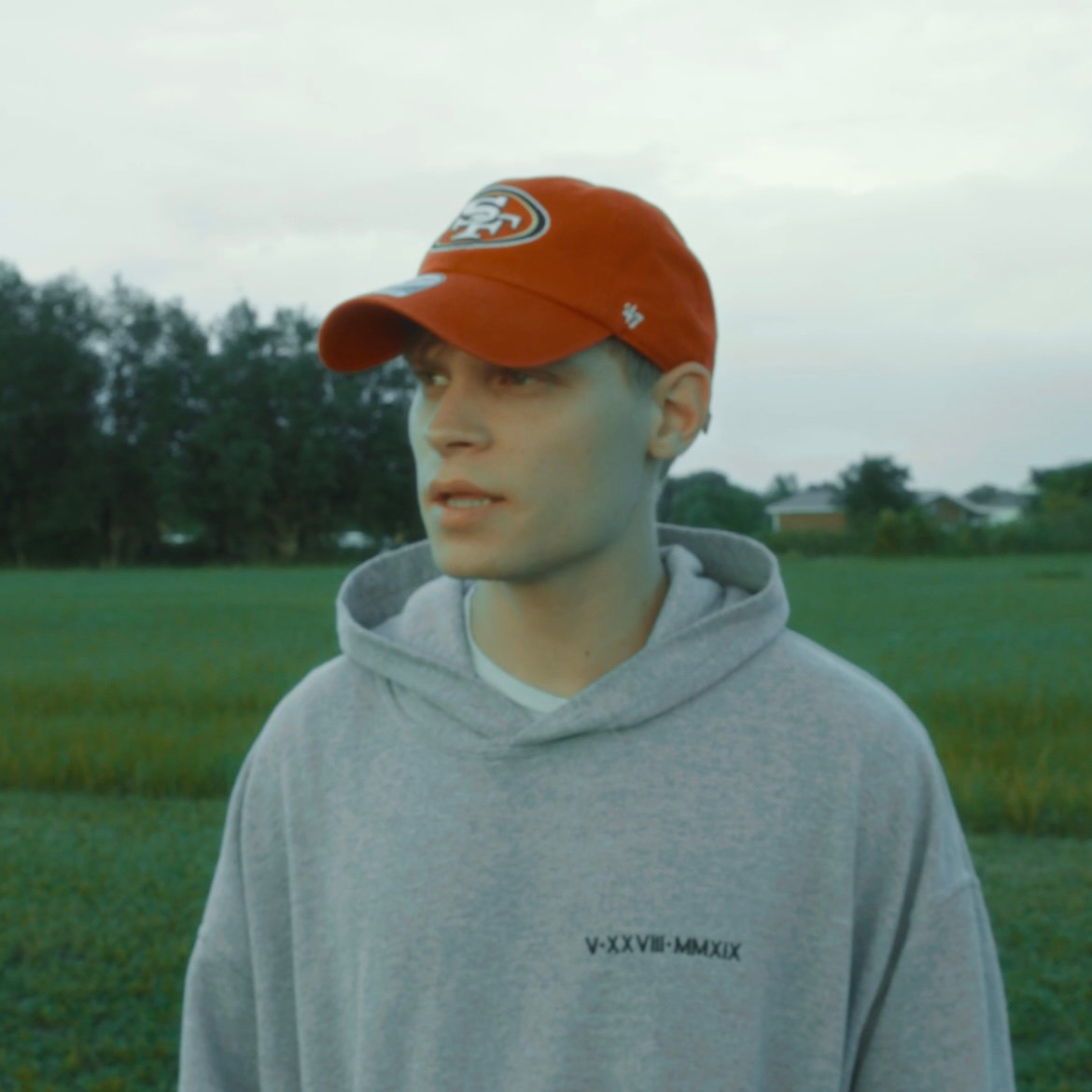 Young man wearing a red San Francisco 49ers cap and gray hoodie standing in a green field with trees in the background.
