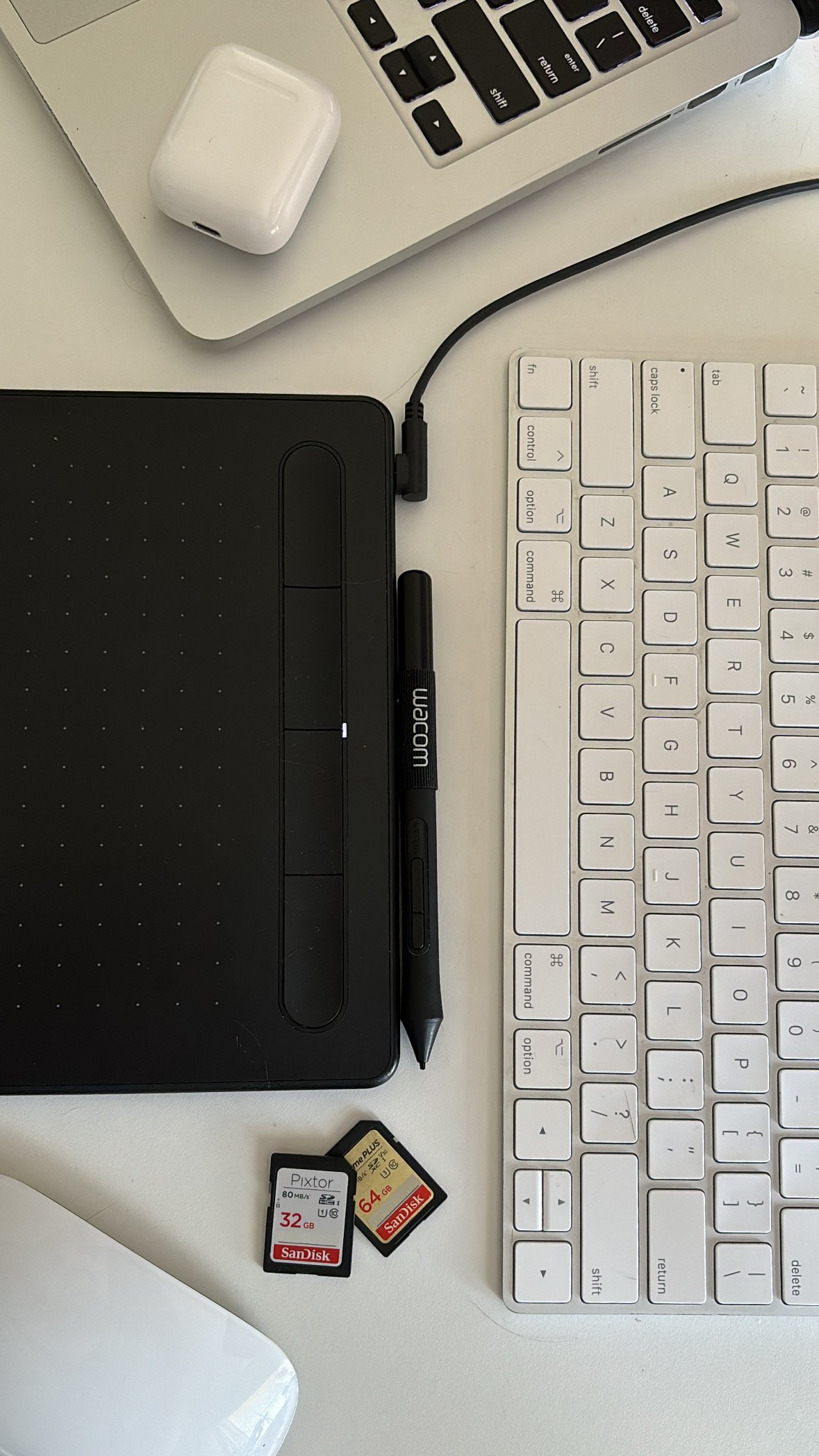 Workspace with a Wacom drawing tablet and pen, white keyboard, AirPods case, white mouse, and two SD memory cards on a white surface.