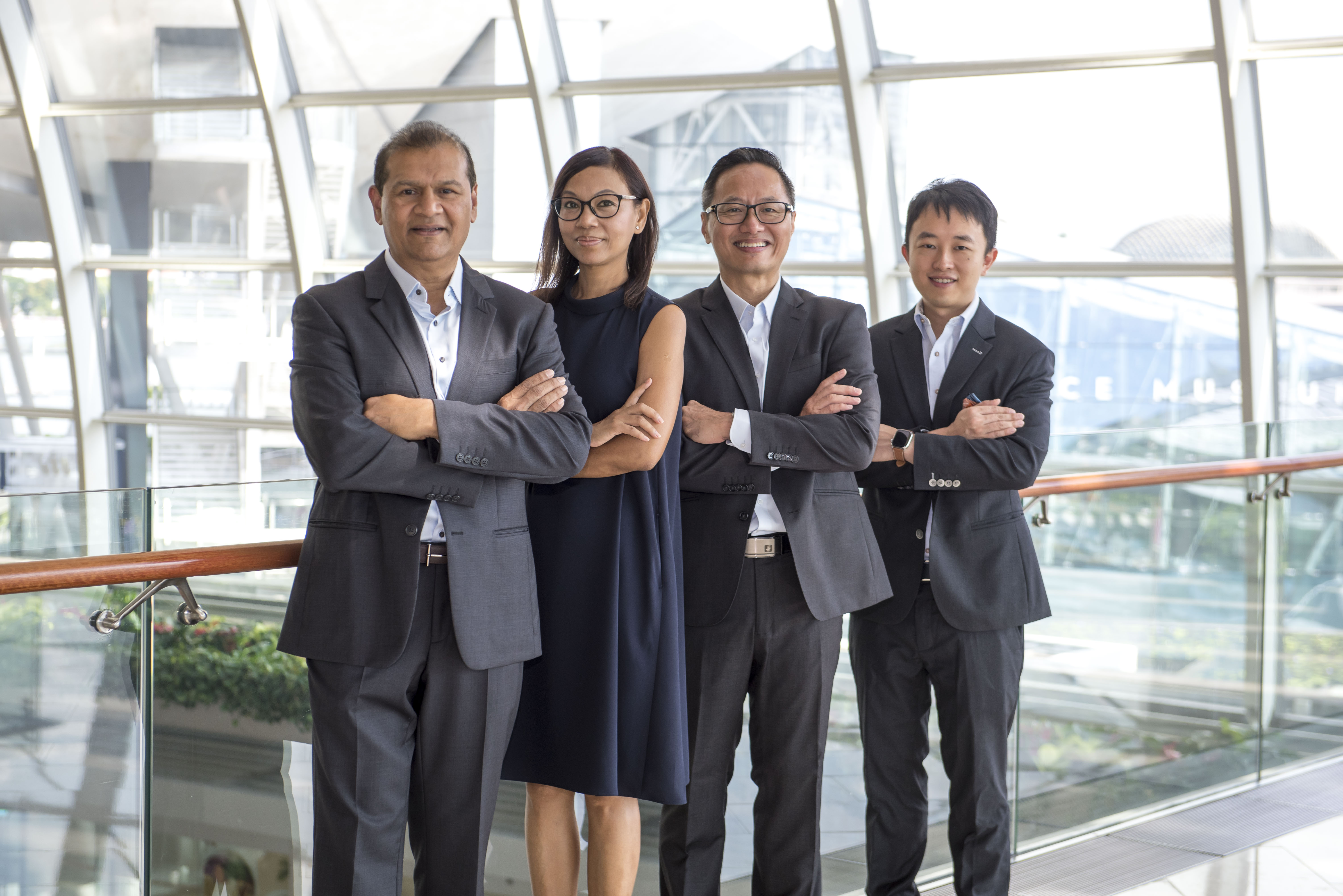 Four business professionals standing indoors in front of large windows with arms crossed, smiling at the camera; three men in dark suits and one woman in a sleeveless dark dress.