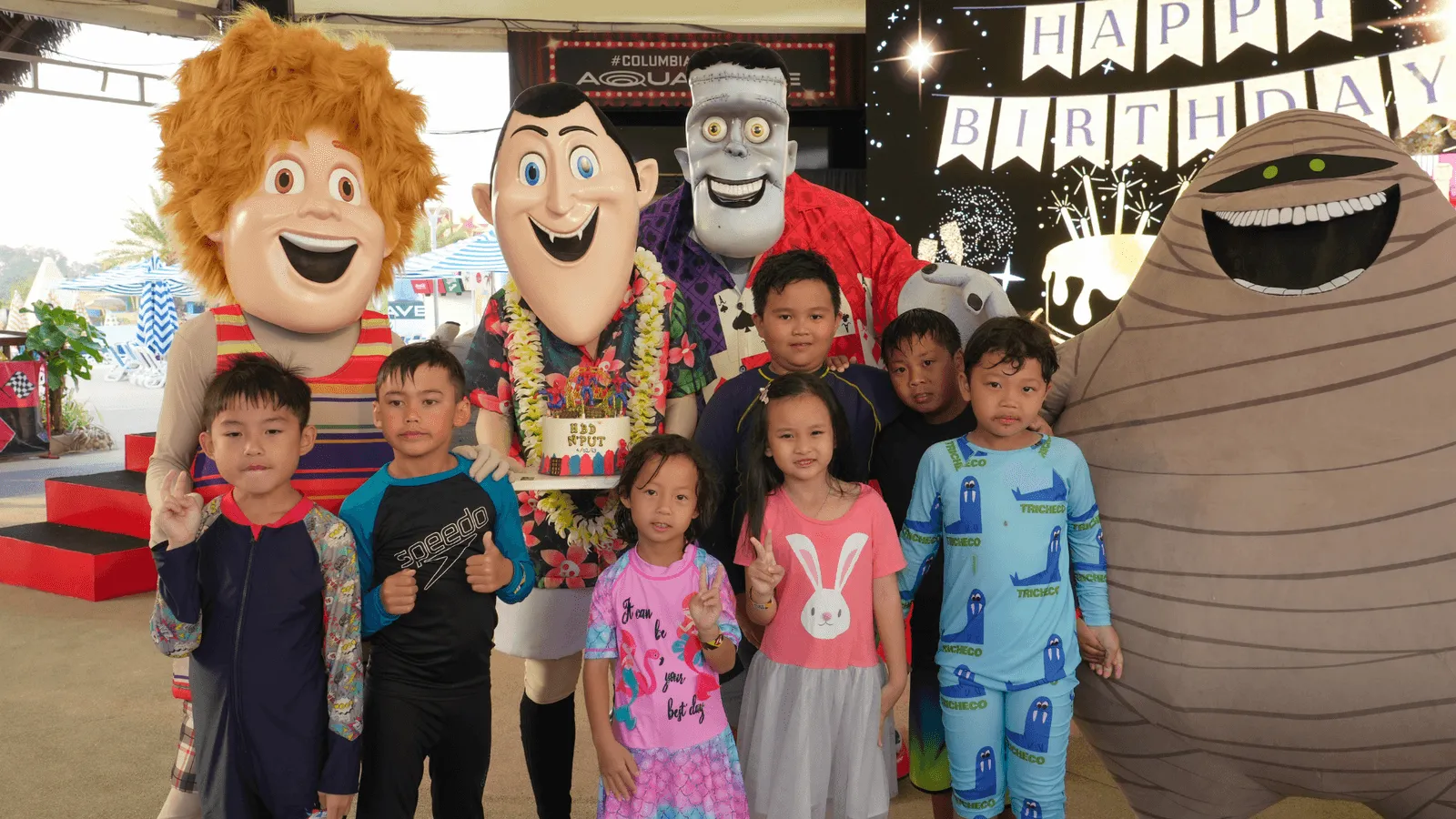 A group of smiling children poses with life-sized mascots from Hotel Transylvania, including Johnny, Dracula, Frankenstein, Murray the mummy, and the werewolf. The kids, dressed in colorful pajamas and casual clothes, stand in front of a birthday-themed stage decorated with a large “HAPPY BIRTHDAY” banner and festive lights. The Dracula mascot holds a birthday cake prop, adding to the celebratory atmosphere. The scene is lively, joyful, and perfect for a themed birthday party at Columbia Pictures Aquaverse.