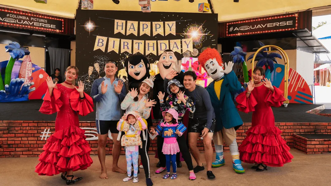 A family poses happily with Hotel Transylvania character mascots and dancers at a themed birthday celebration at Columbia Pictures Aquaverse. The background features a festive stage decorated with a large "HAPPY BIRTHDAY" banner, colorful props, and cartoon backdrops. The group smiles and waves at the camera, surrounded by performers in red flamenco-style dresses and costumed characters like Dracula, Mavis, and Johnny. The atmosphere is cheerful and celebratory, perfect for a fun-filled birthday event at the park.