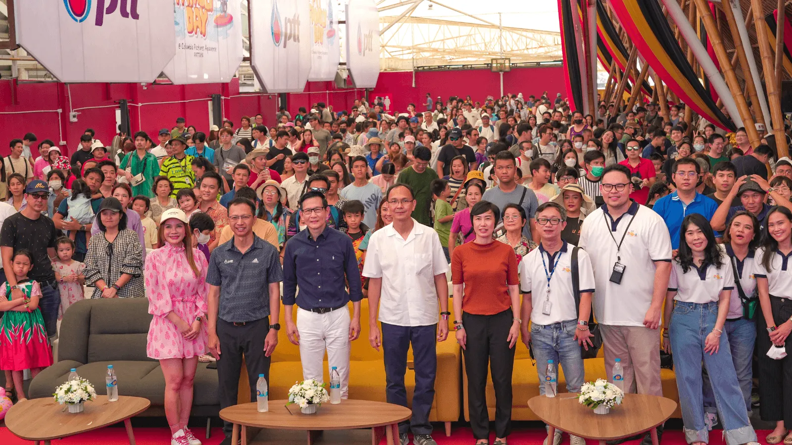 A large crowd gathers indoors at a brightly decorated event venue. In the foreground, a group of event hosts and organizers—six men and three women—stand smiling for the camera in front of two round tables with white flower arrangements. Behind them, hundreds of attendees fill the space, creating a festive and energetic atmosphere. The venue is adorned with colorful hanging decorations and banners, and the lighting is warm and inviting. Everyone appears to be enjoying a formal or celebratory occasion.