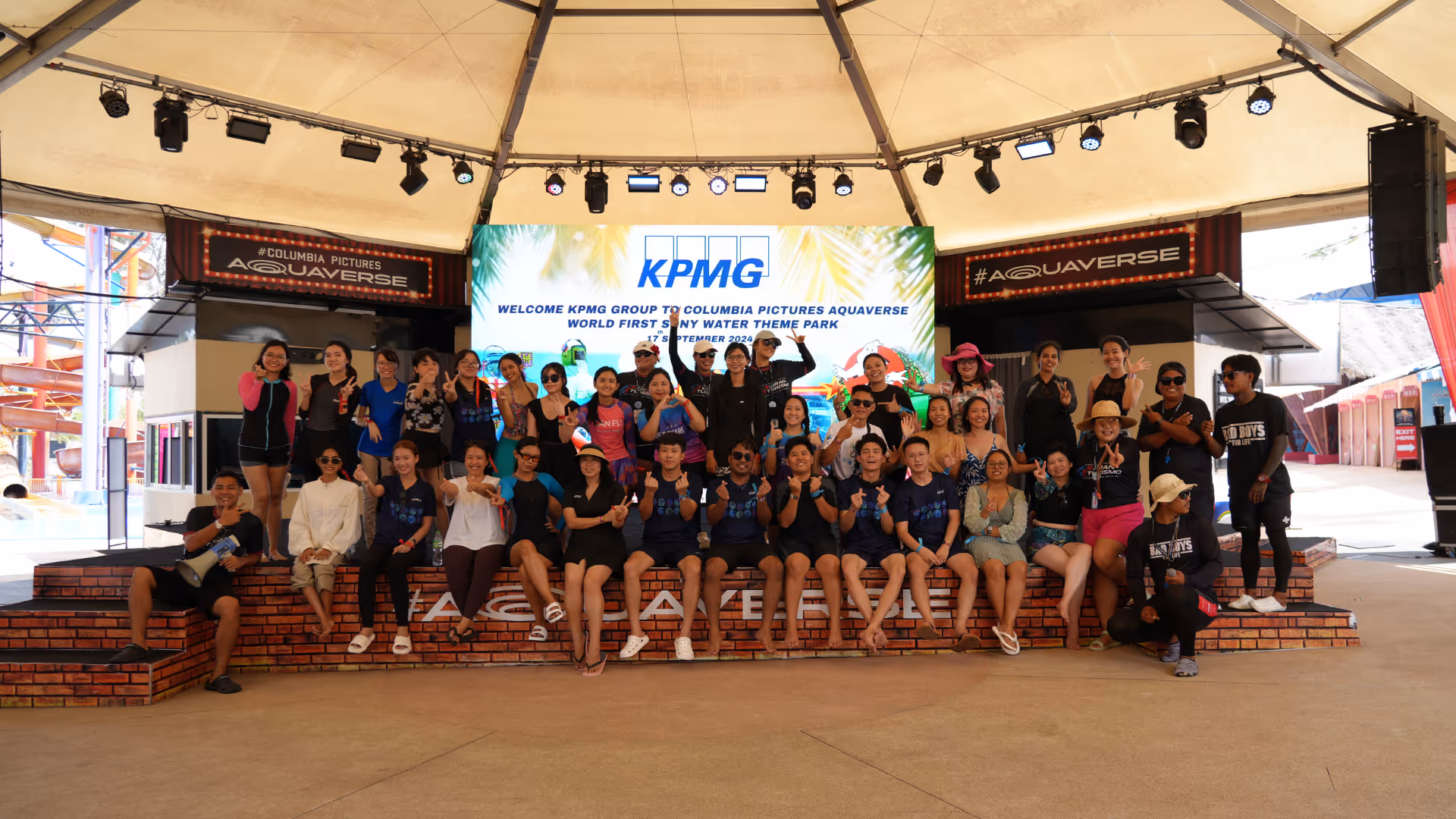 A large group of people pose together on a stage under a circular open-air pavilion with a high ceiling. Behind them is a large digital screen displaying the KPMG logo and the text: “KPMG ASEAN and South Asia Future Leaders Summit.” The group, consisting of around 50 individuals, is smiling, laughing, and making celebratory gestures, indicating a fun and energetic atmosphere. The venue appears to be part of a theme park or entertainment complex, with amusement rides and signage partially visible in the background.


