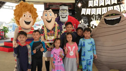 A group of young children in colorful pajamas and swimsuits pose for a photo with life-sized mascots from the animated movie Hotel Transylvania. The characters include Johnny, Dracula, Frankenstein, Murray the mummy, and a red-suited werewolf, all smiling cheerfully. Behind them is a festive backdrop with a “Happy Birthday” banner, balloons, and decorations, indicating a birthday celebration. The children are smiling, making peace signs, and standing confidently in front of the characters, creating a joyful and vibrant scene.