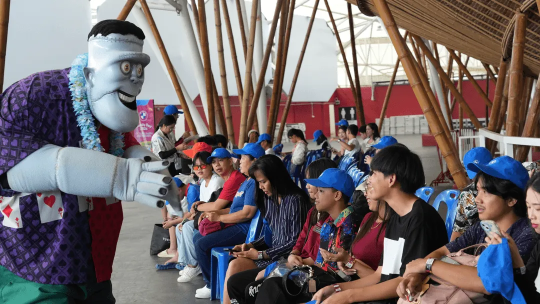 A friendly Frankenstein mascot from Hotel Transylvania, dressed in a purple shirt and wearing a Hawaiian lei, interacts playfully with seated guests at Columbia Pictures Aquaverse. The guests, many of whom are wearing matching blue caps, smile and engage with the character. The setting is shaded with bamboo beams and open space, suggesting an organized group visit or corporate outing. The mood is cheerful and lighthearted, with the mascot bringing entertainment and laughter to the group.