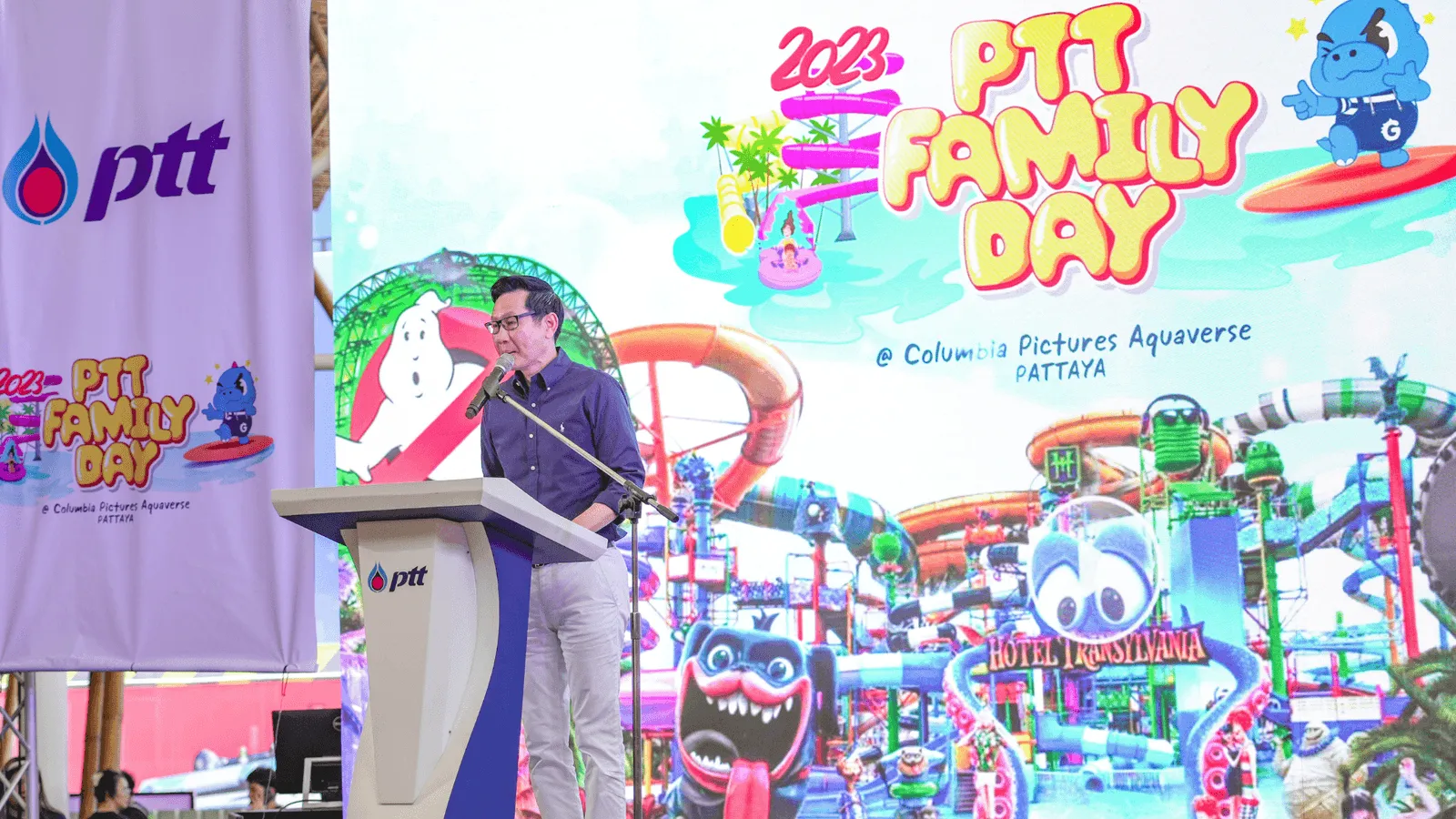 A man in formal attire stands at a podium delivering a speech during the 2023 PTT Family Day event at Columbia Pictures Aquaverse. Behind him, a large, colorful screen displays the event’s branding, including vibrant water slides and cartoon characters from the Hotel Transylvania attraction. Large PTT banners flank the stage, emphasizing the corporate sponsorship. The setting is bright and festive, capturing the excitement of the event’s opening ceremony or keynote moment.