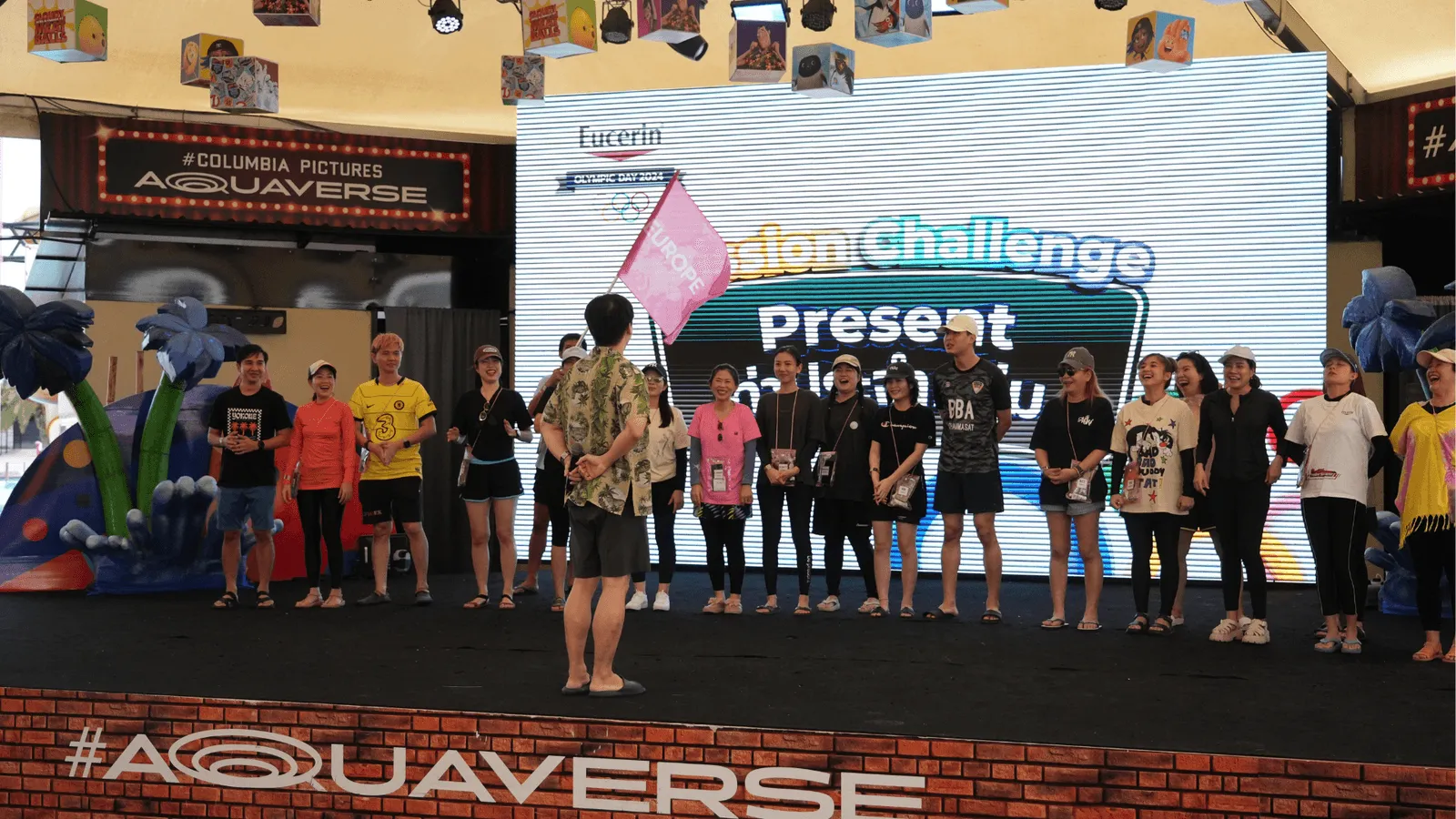 A group of participants stand on an indoor stage at Aquaverse, facing a host in a floral shirt holding a pink flag. The backdrop features a large screen displaying the text “Mission Challenge Present,” with colorful graphics and branding. The participants wear casual or sporty attire, including t-shirts, caps, and shorts, suggesting involvement in a team activity or game show. The stage is surrounded by fun cartoon props and banners, creating a lively, energetic atmosphere typical of a themed competition or event.