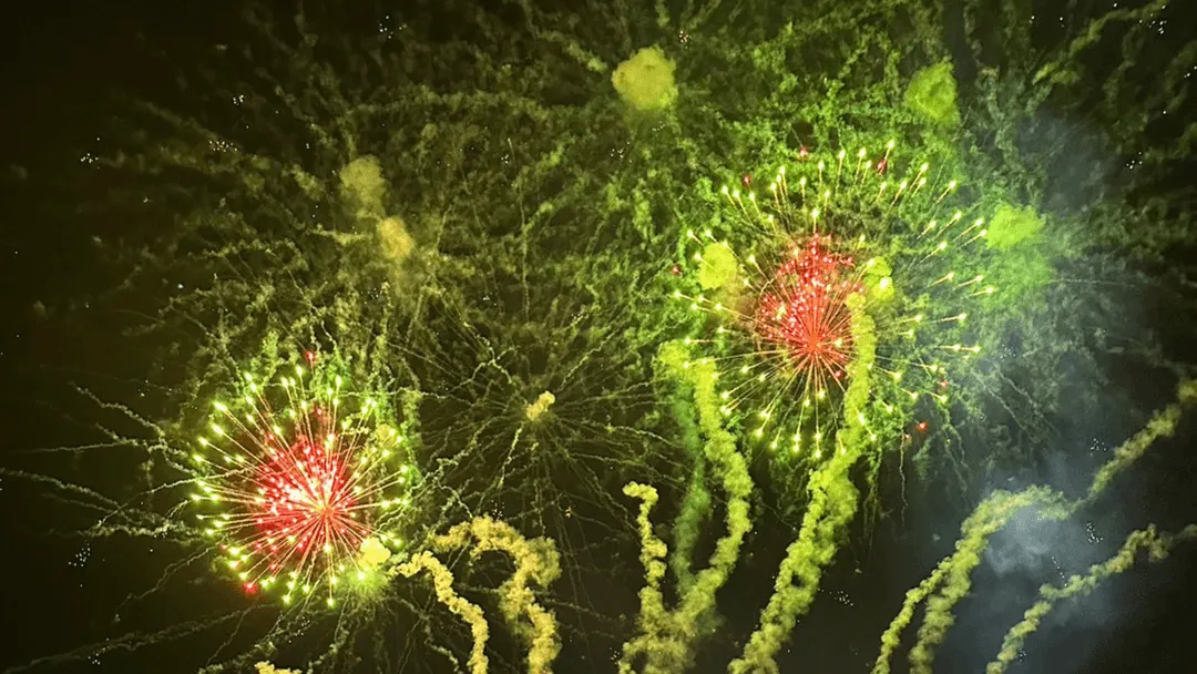 A vibrant fireworks display lights up the night sky with dazzling bursts of green and red. Three large firework explosions radiate outward like glowing flowers, surrounded by swirling trails of smoke and sparks. The bright colors contrast sharply against the dark background, creating a dramatic and celebratory scene filled with energy and motion.