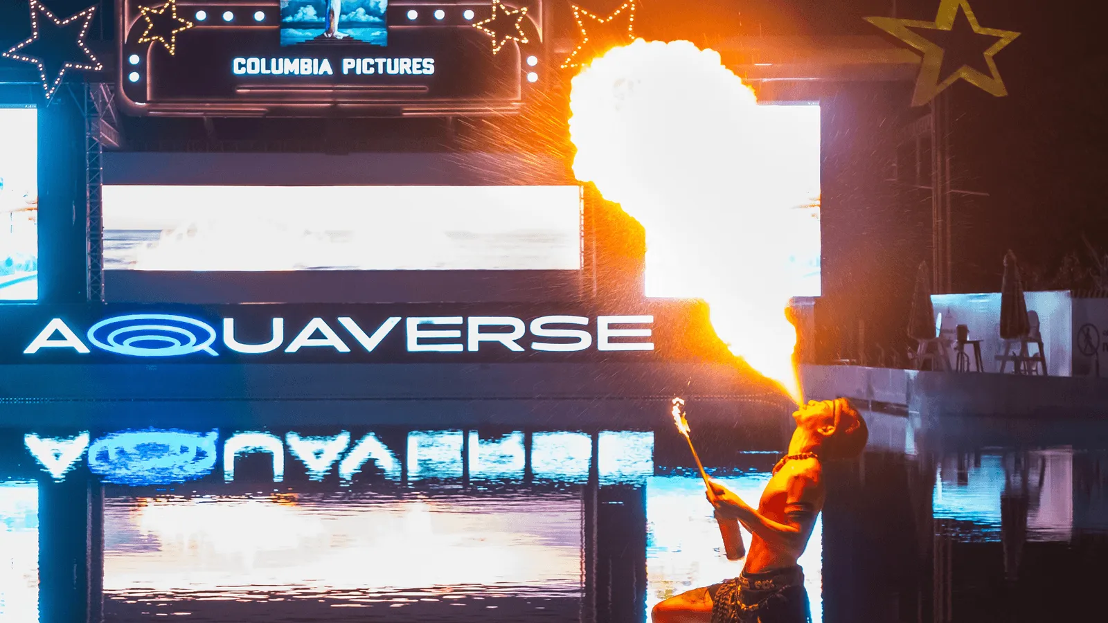 A dramatic night performance at Columbia Pictures Aquaverse features a fire breather exhaling a massive plume of flame into the air. The performer stands by the water's edge, with bright signage reading “AQUAVERSE” and “COLUMBIA PICTURES” illuminated behind them and reflecting on the water below. Golden star decorations add to the festive ambiance, enhancing the high-energy, theatrical vibe of the event.