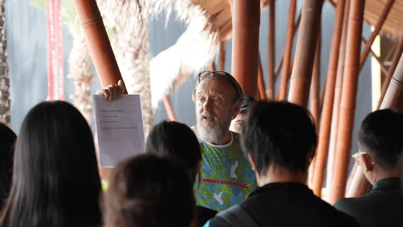 An older man with a gray beard and colorful sweater addresses a small group of people, holding up a printed sheet of paper. He appears to be giving instructions or leading a discussion, with his audience standing attentively in front of him. The setting includes tall wooden beams and tropical plants, suggesting an outdoor or semi-outdoor environment, possibly part of a cultural or educational tour. Sunlight filters through, creating a warm, natural atmosphere.