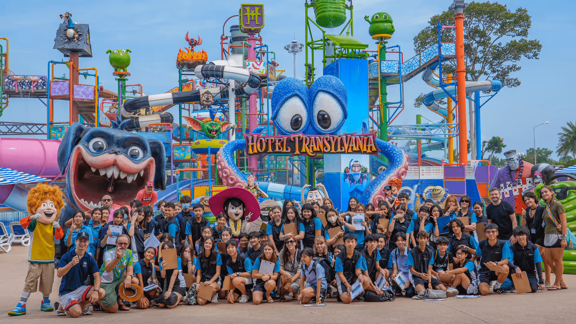 A large group of young people, many holding guitars, pose in front of the vibrant and colorful Hotel Transylvania water play zone at a theme park. Behind them are oversized cartoon characters, water slides, splash towers, and playful decorations featuring characters from the Hotel Transylvania movie franchise. Mascots of Johnny and other characters stand with the group, enhancing the fun atmosphere. The sky is clear, and the setting is lively, festive, and full of excitement, suggesting a school or group trip to the Aquaverse water park.