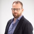 Man with short hair and beard wearing glasses, a checked shirt, and a dark blazer against a plain light background.