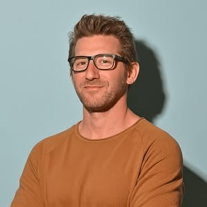 Man with glasses and brown sweater smiling against a light blue background.