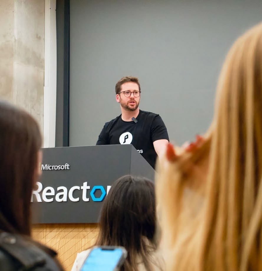 A man with glasses speaking at a Microsoft Reactor event podium, with an audience in the foreground.