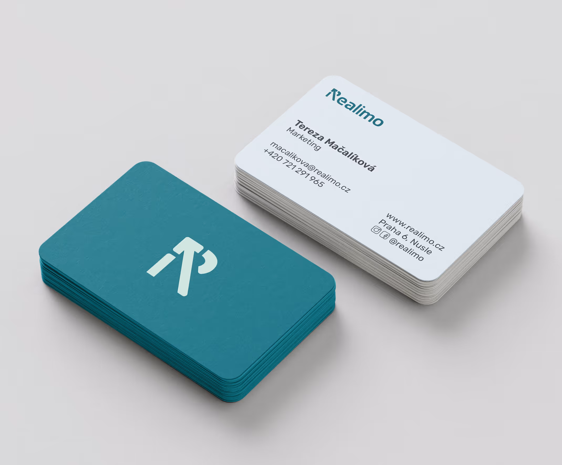 Dva balíčky vizitek na světle šedém pozadí, jedna strana v tyrkysové barvě s logem, druhá strana bílá s kontaktními údaji Terezy Mačalíkové z marketingu společnosti Realimo.
