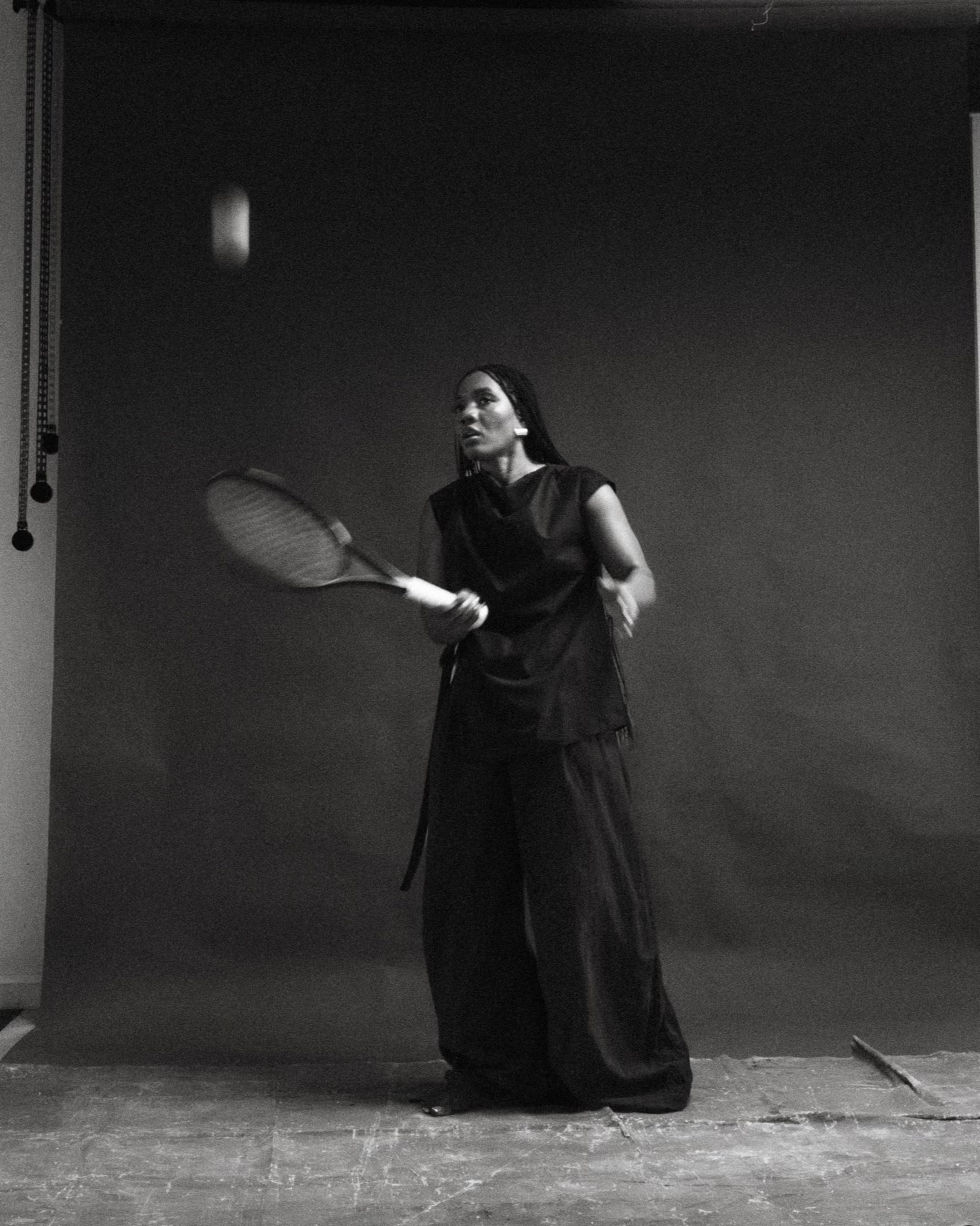 Naa Shika adu B&W photo tennis racket poised to hit a ball against a plain dark backdrop..
