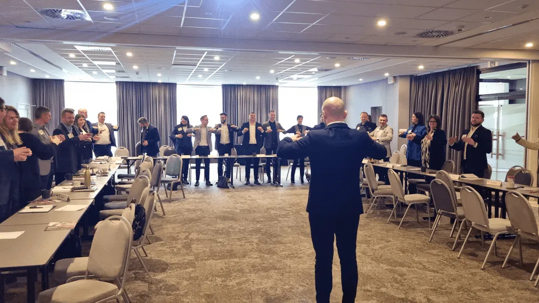 Rafał Żebrowski prowadzi warsztaty w sali konferencyjnej, stojąc przed grupą uczestników, którzy stoją w półokręgu. Obraz ilustruje jego misję pomagania ludziom w rozwijaniu umiejętności komunikacyjnych i przywódczych.