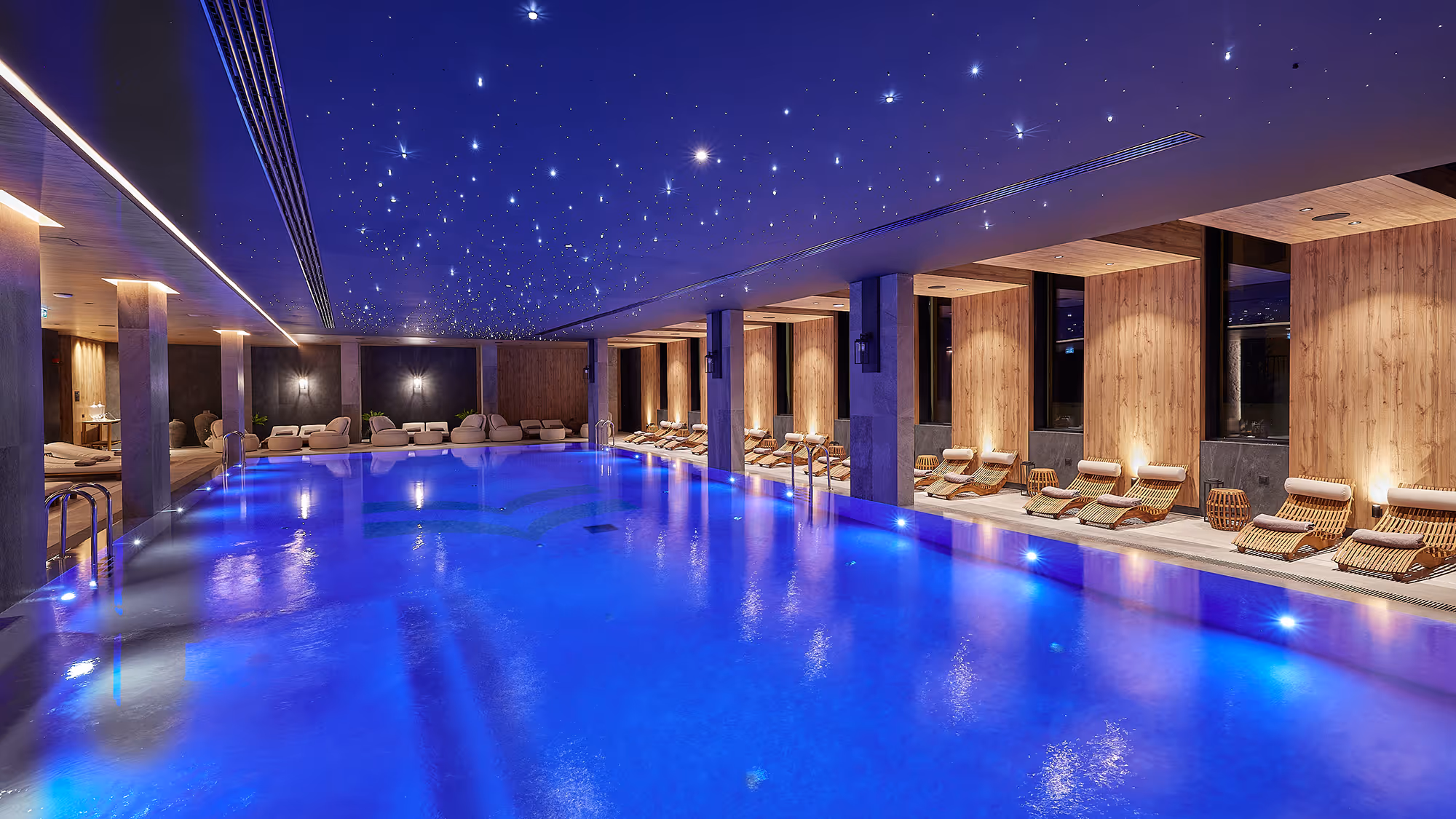 Indoor swimming pool lit with blue lights, surrounded by wooden lounge chairs and ceiling lights resembling stars.