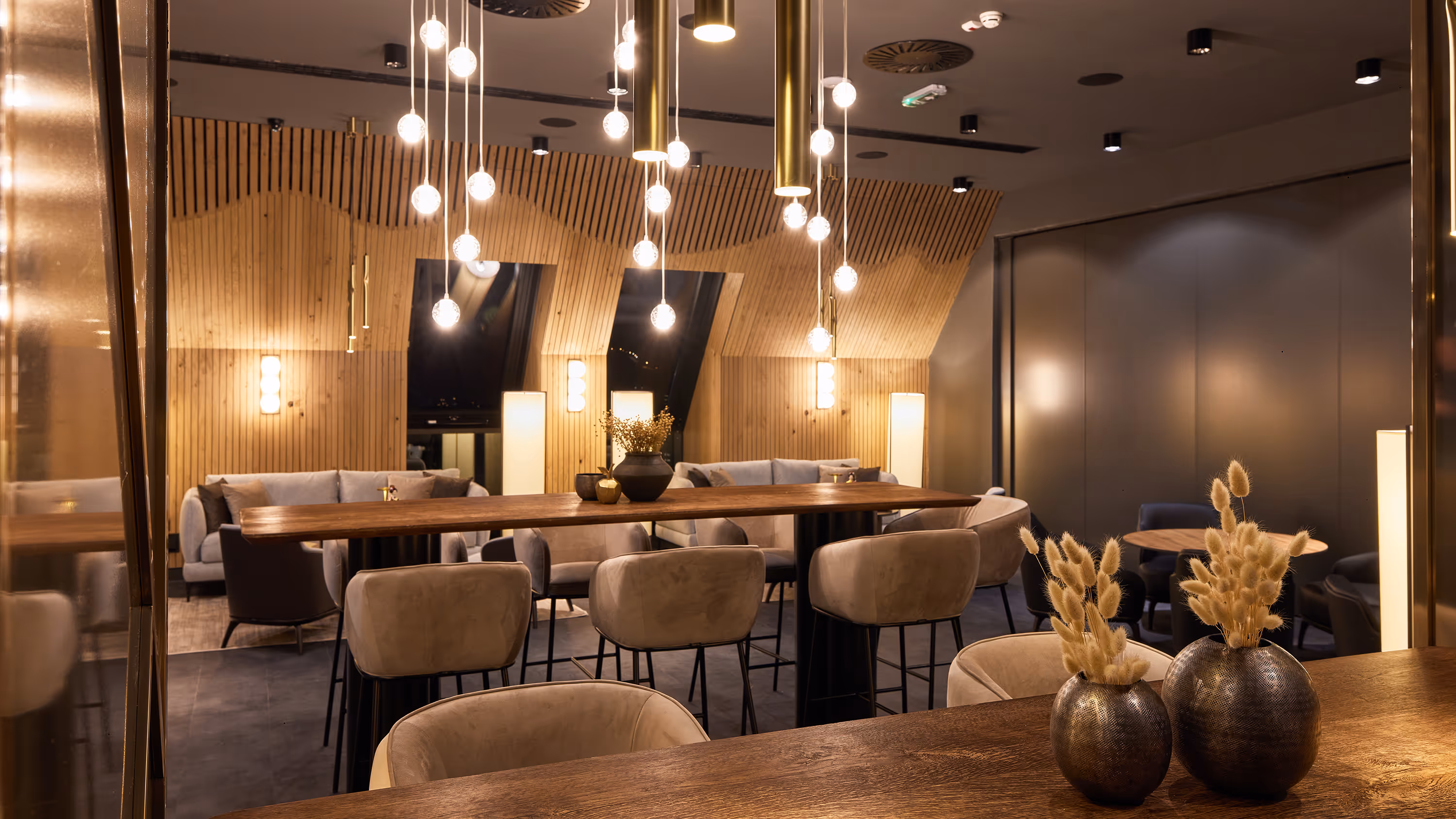 Modern lounge area with wooden tables, beige chairs, hanging pendant lights, and decorative vases with dried plants.