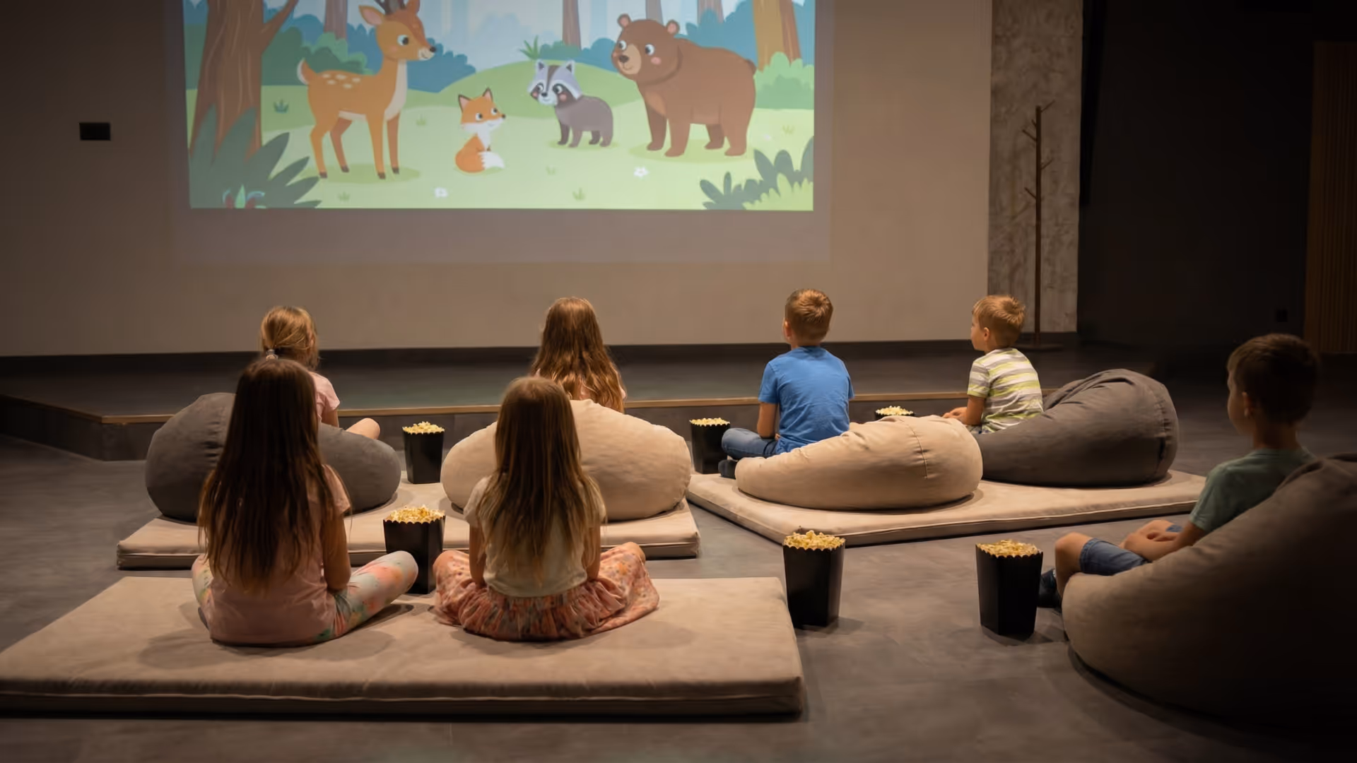 Seven children sitting on cushions and bean bags watching a nature-themed cartoon projected on a wall.