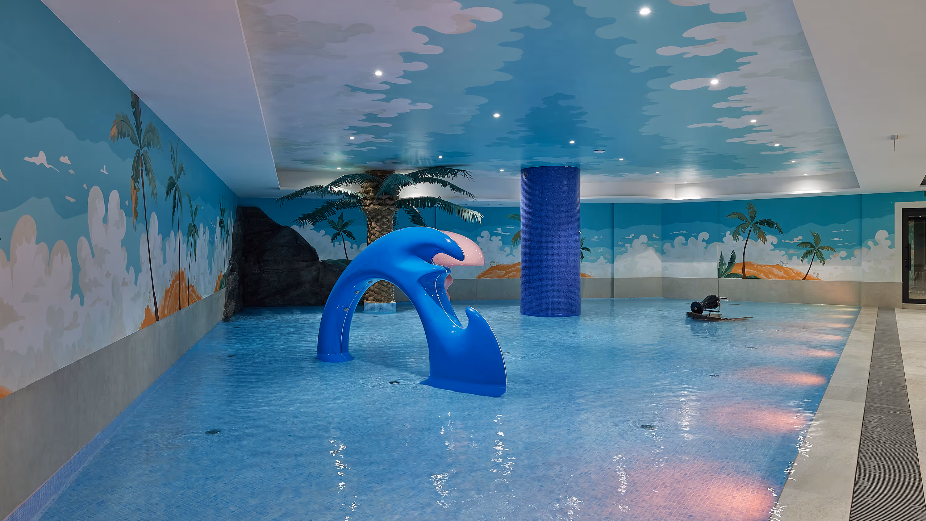 Indoor children's pool with blue water, a blue whale-themed slide, palm tree decorations, and a painted tropical mural on the walls and ceiling.