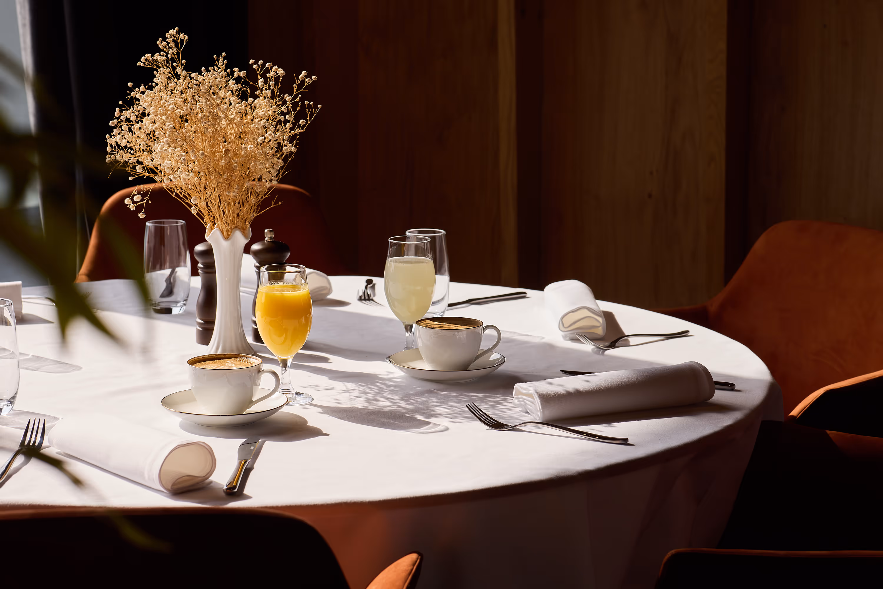 Round dining table with white tablecloth, two cups of coffee, two glasses of juice, white napkins, and a vase of dried flowers.