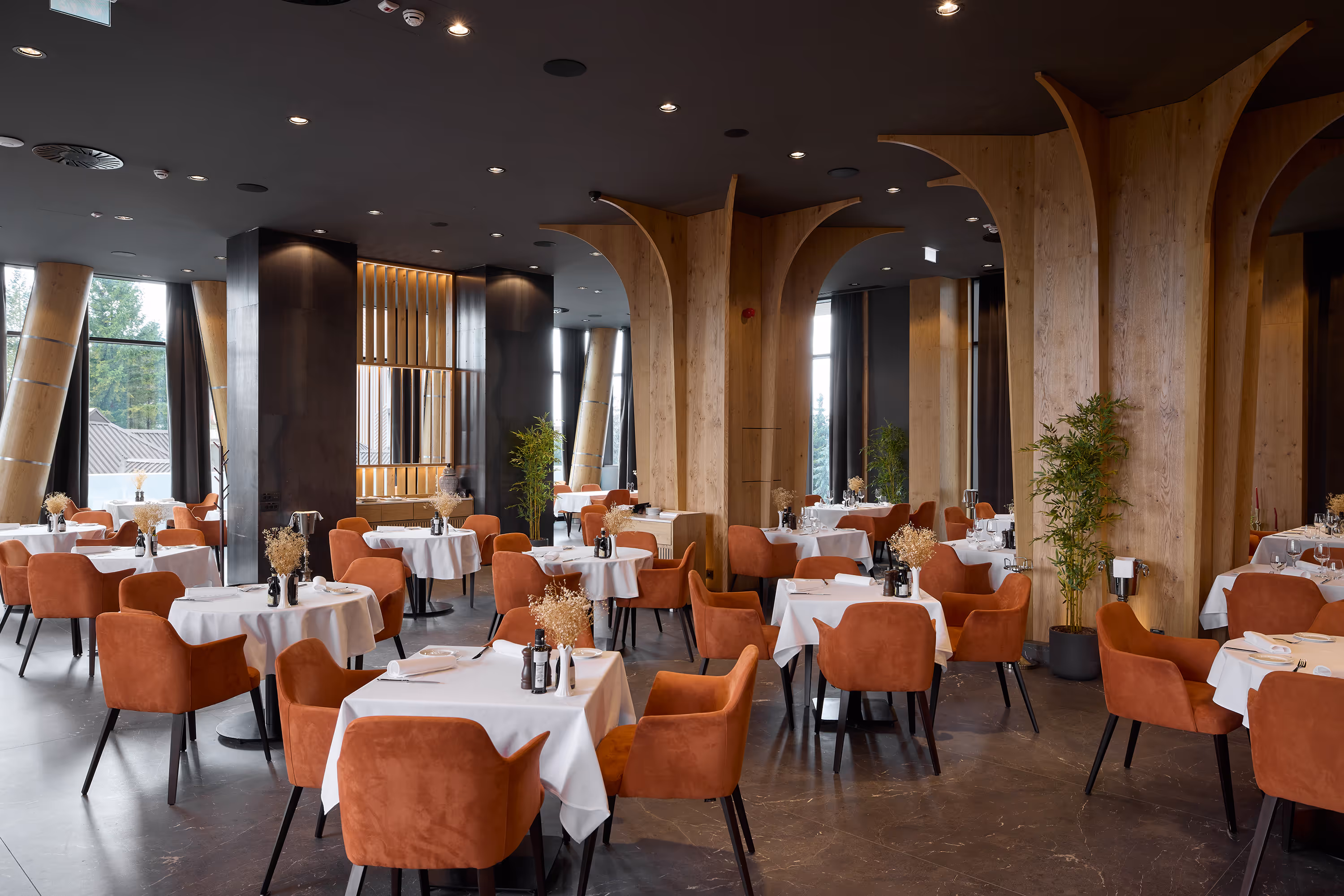 Modern restaurant interior with round and square tables covered in white tablecloths, orange upholstered chairs, wooden columns, and large windows.