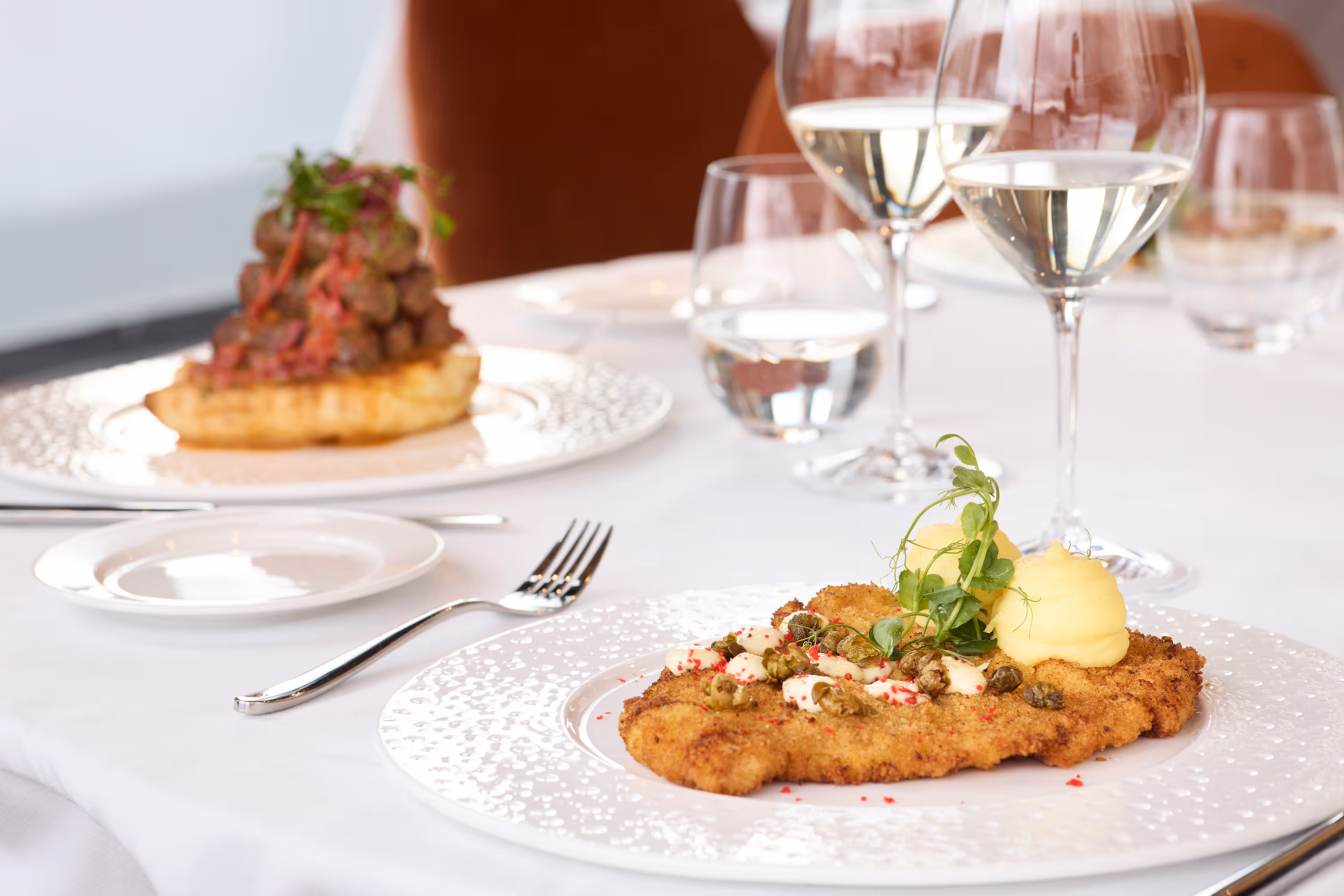 Breaded schnitzel garnished with mashed potatoes, capers, and microgreens on a white patterned plate with wine glasses and another dish in the blurred background.