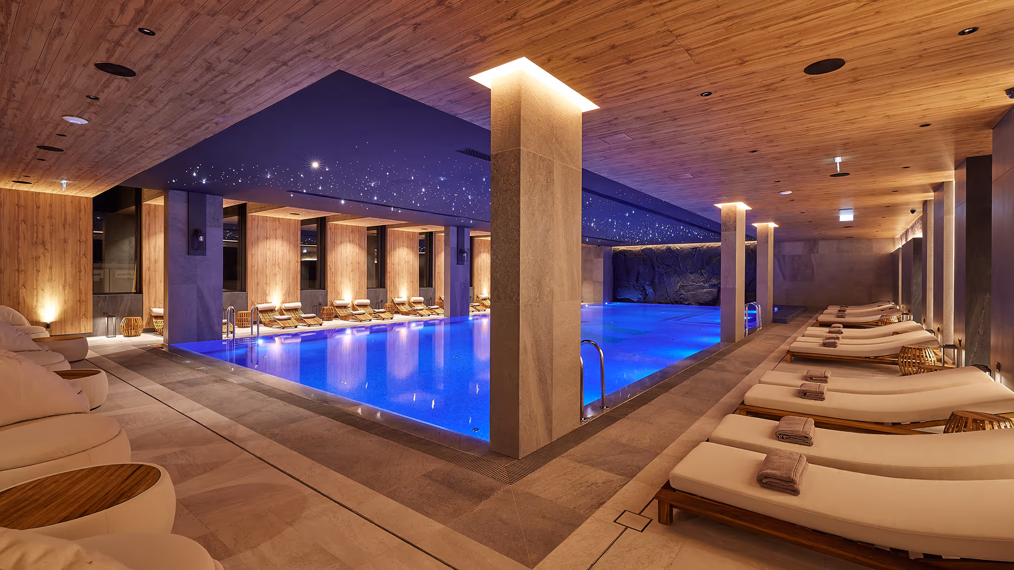 Indoor spa pool with blue lighting, wooden ceiling, stone columns, and cushioned lounge chairs with towels.