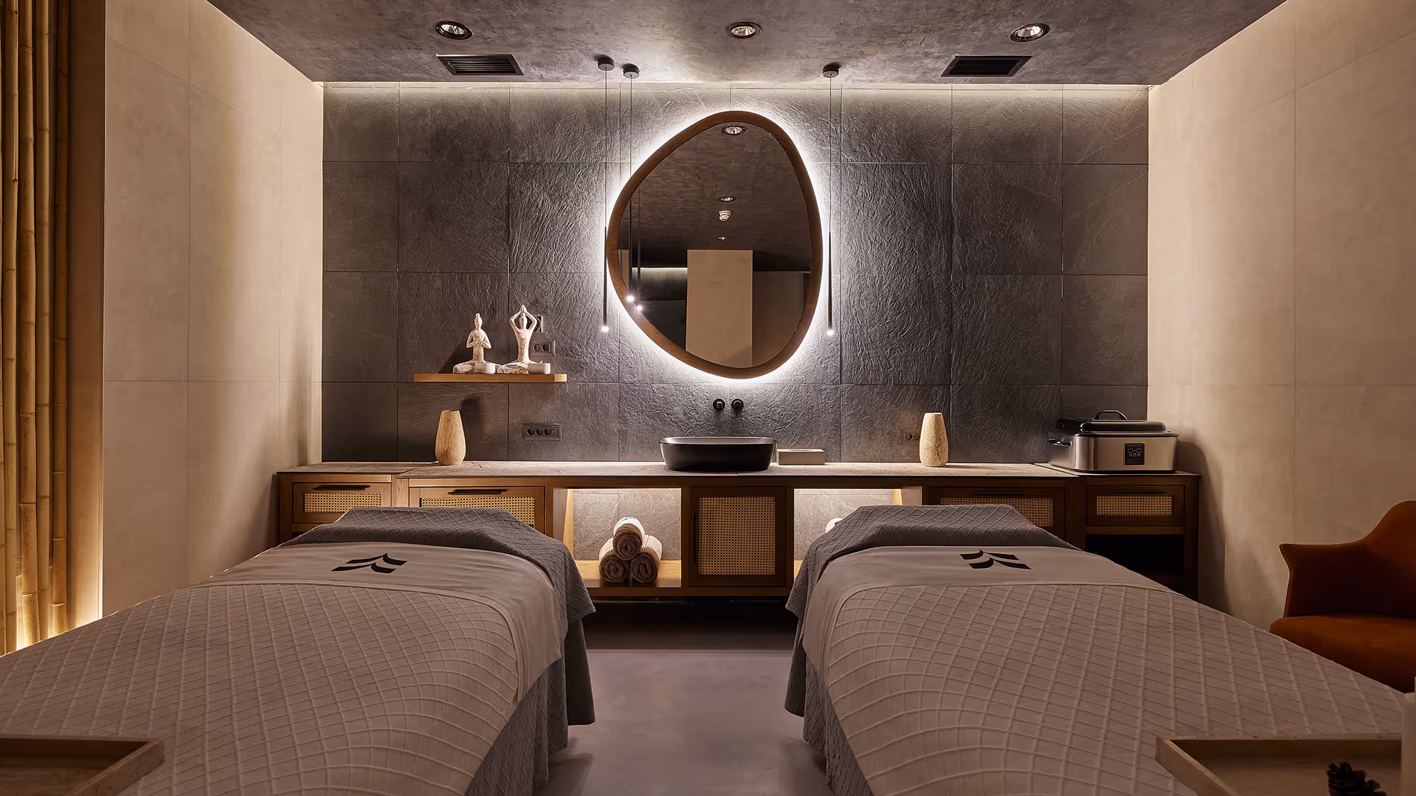 Modern spa room with two massage tables, illuminated irregularly shaped mirror, and calming neutral tones.