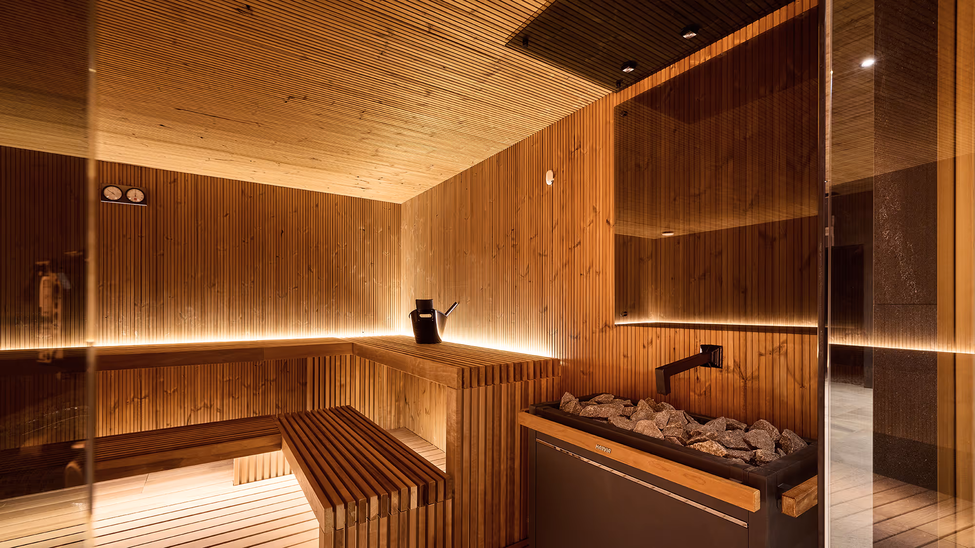 Interior of a modern wooden sauna with benches, a bucket with a ladle, and heater with rocks.