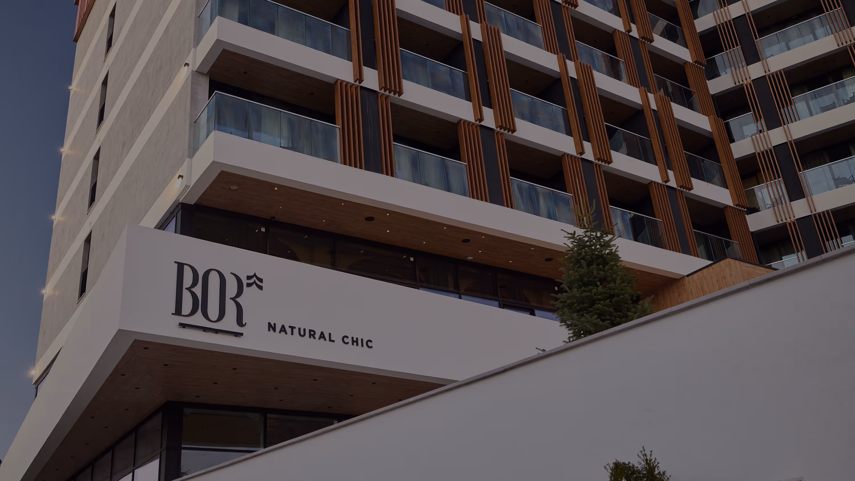 Modern building facade with balconies, featuring a sign reading 'BOR NATURAL CHIC' and a small evergreen tree near the entrance.