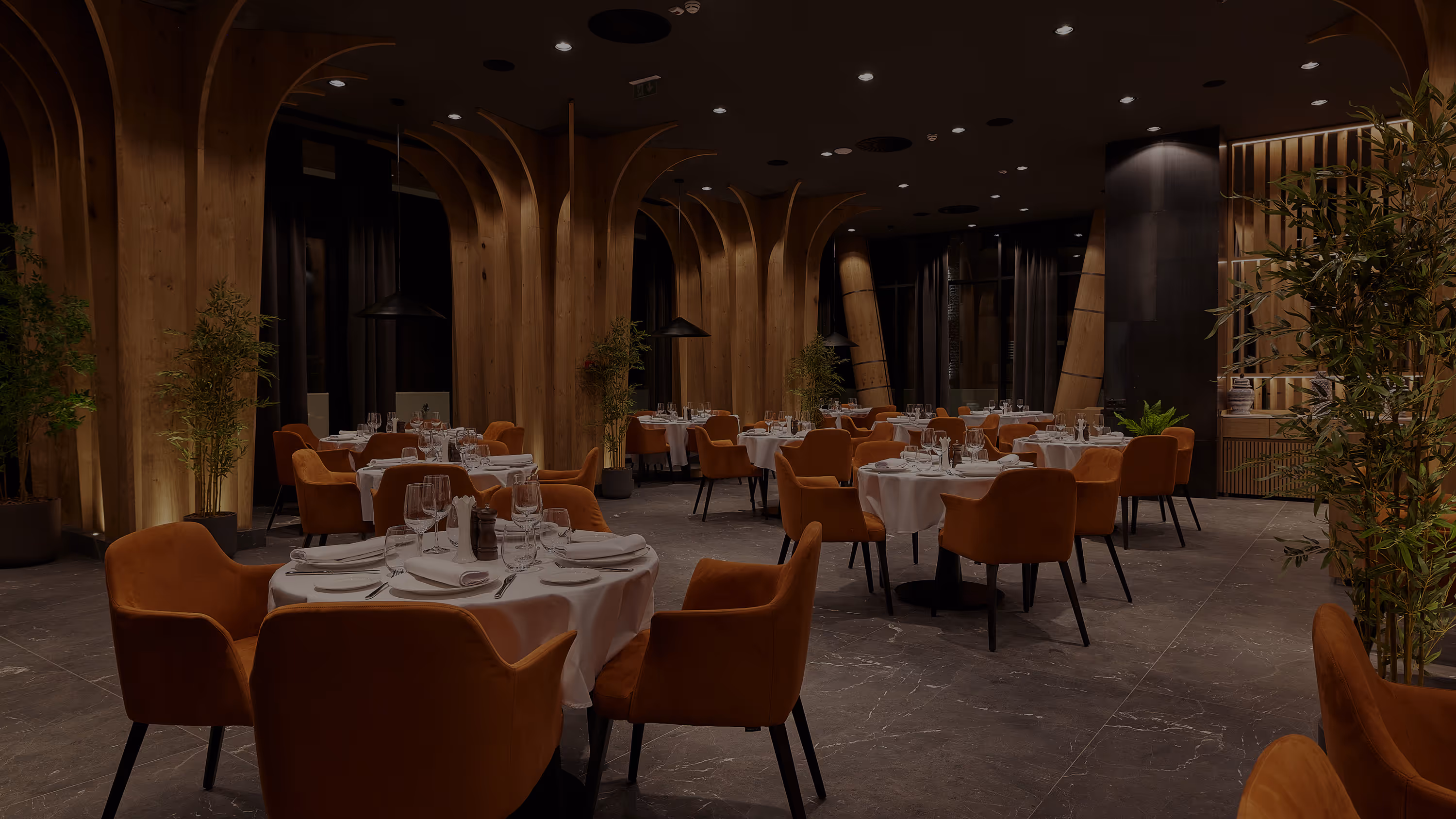 Elegant restaurant interior with round tables set with white tablecloths, orange chairs, and decorative wooden arches.