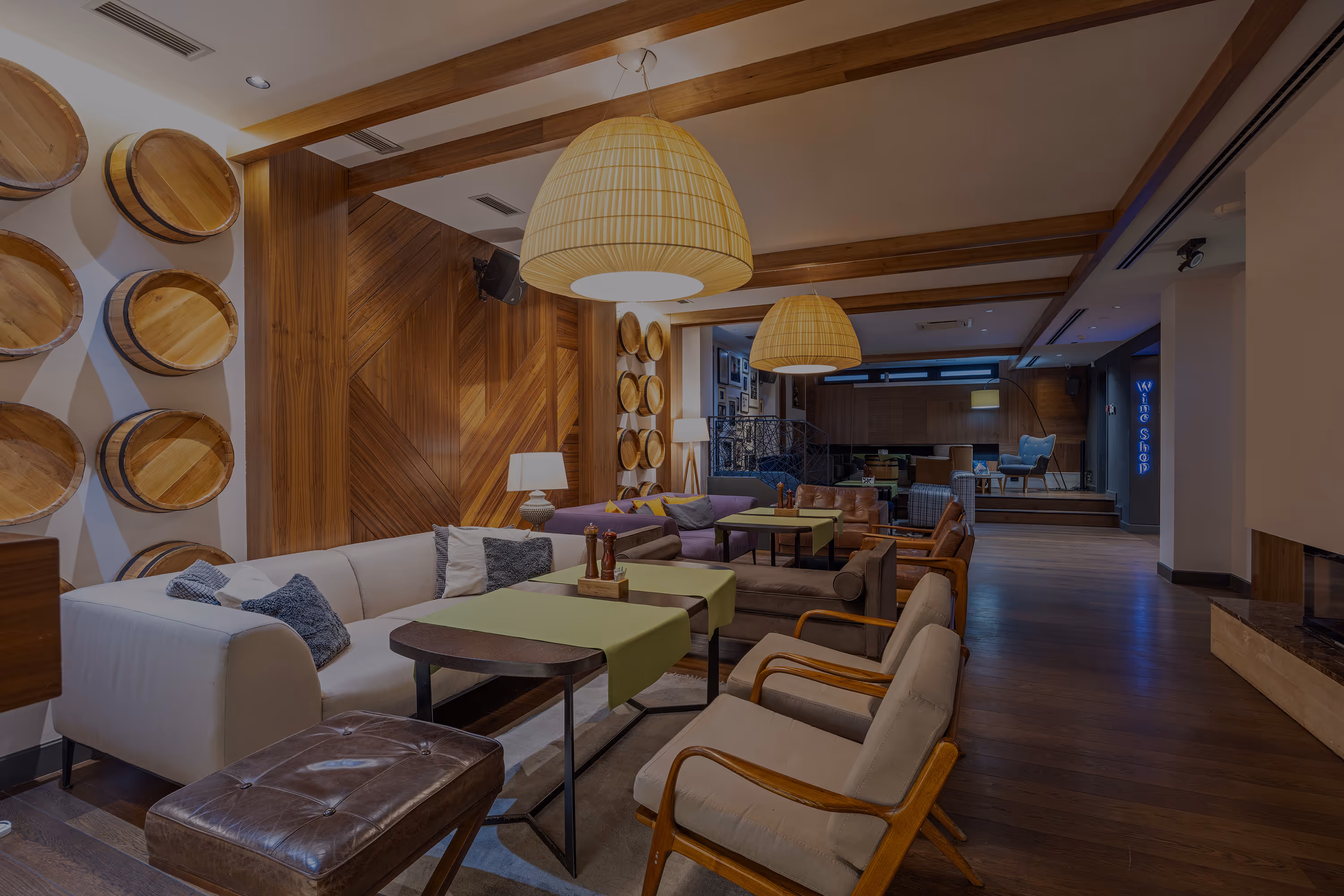 Modern lounge area with wooden wall panels, barrel decorations, beige and brown sofas, green table runners, and hanging woven pendant lights.