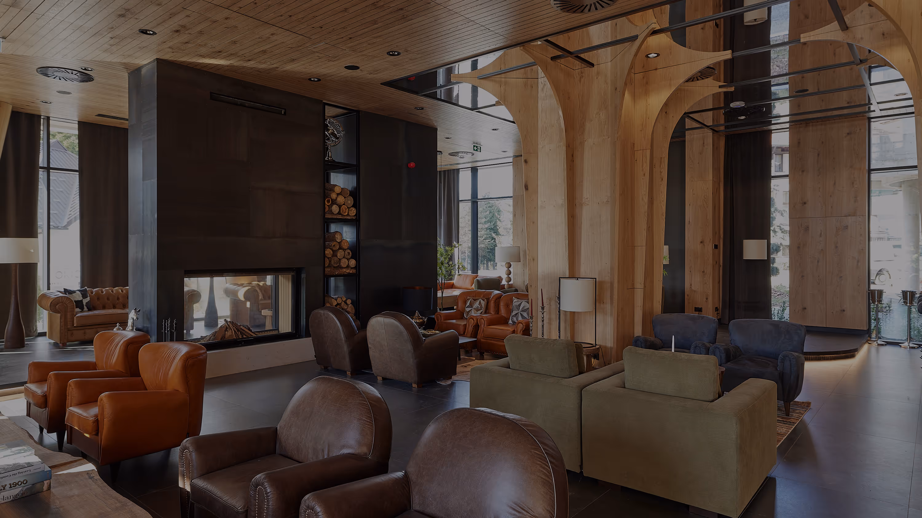 Modern lounge area with various leather and fabric armchairs arranged around a central fireplace with stacked firewood, wooden columns, and large windows.