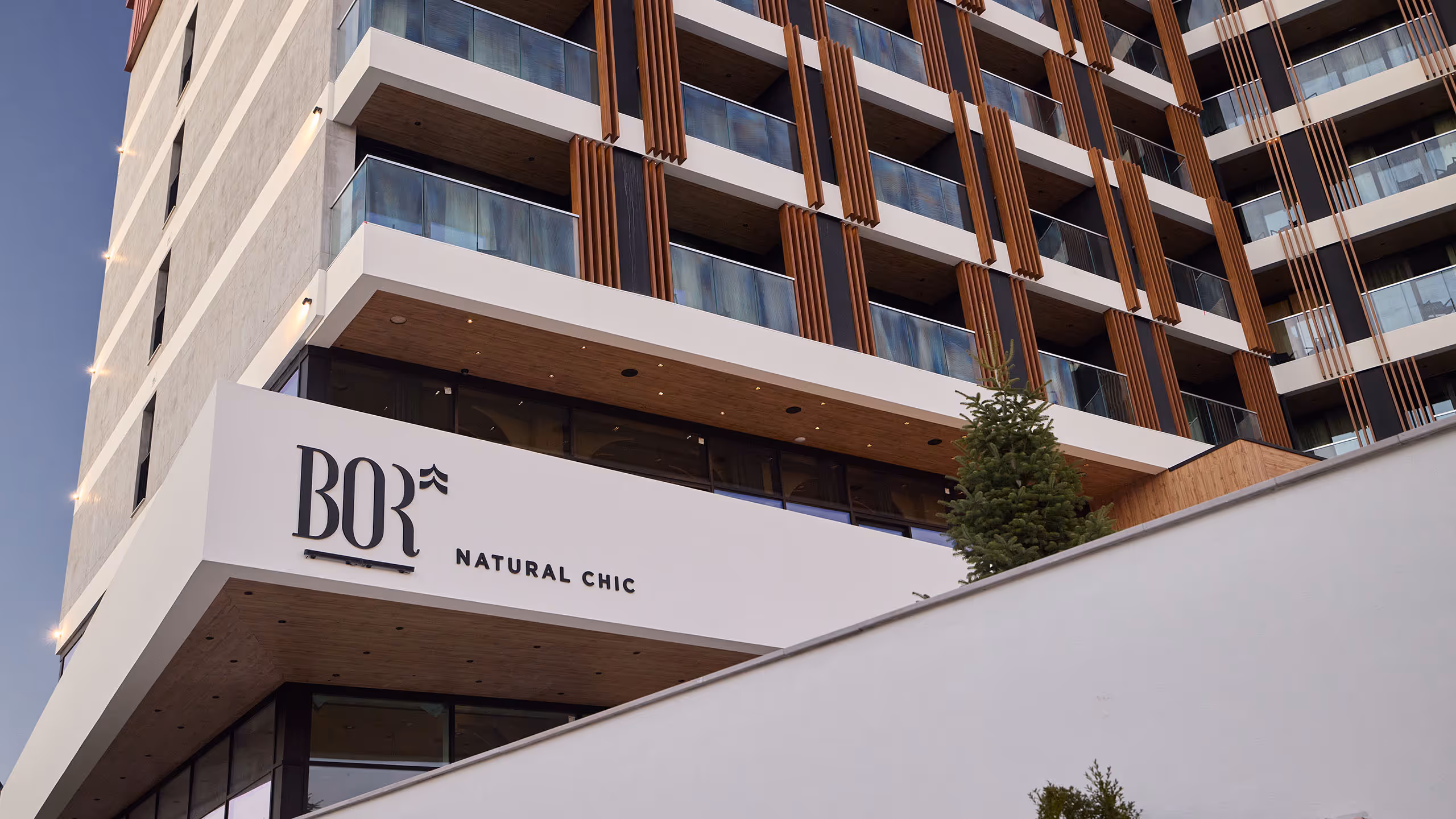 Modern exterior view of Bor Hotel featuring contemporary architecture with glass balconies, natural wood accents, and the Bor Natural Chic logo on the facade.