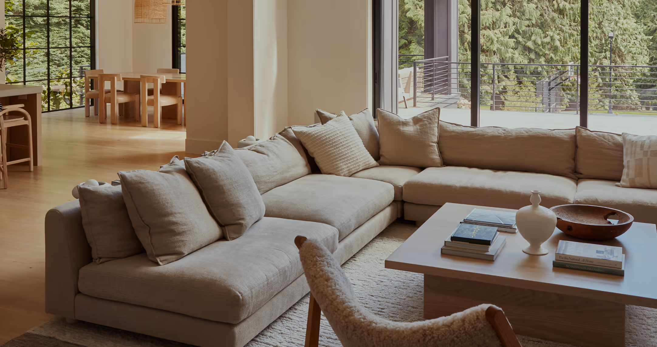 Neutral-toned living room with a large beige sectional sofa, a wooden coffee table with books and decor, and a window showing green trees outside.