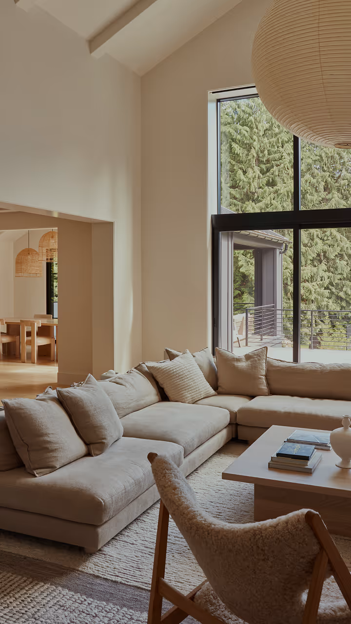 Neutral-toned living room with a large beige sectional sofa, a wooden coffee table with books and decor, and a window showing green trees outside.