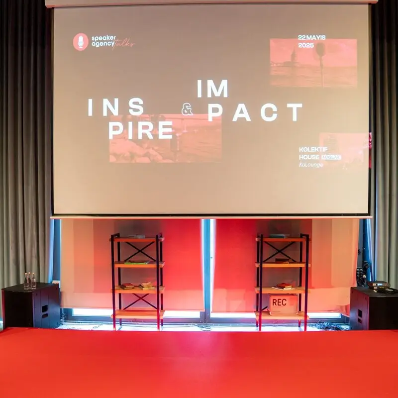 Kadın bir konuşmacı kırmızı halı üzerine oturmuş izleyicilere hitap ediyor, arka planda 'INSPIRE & IMPACT' yazısı görünen sunum ekranı var.