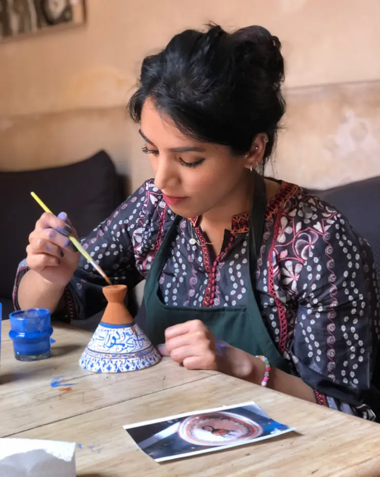Woman painting intricate blue and white patterns on the top of a small ceramic tagine pot.