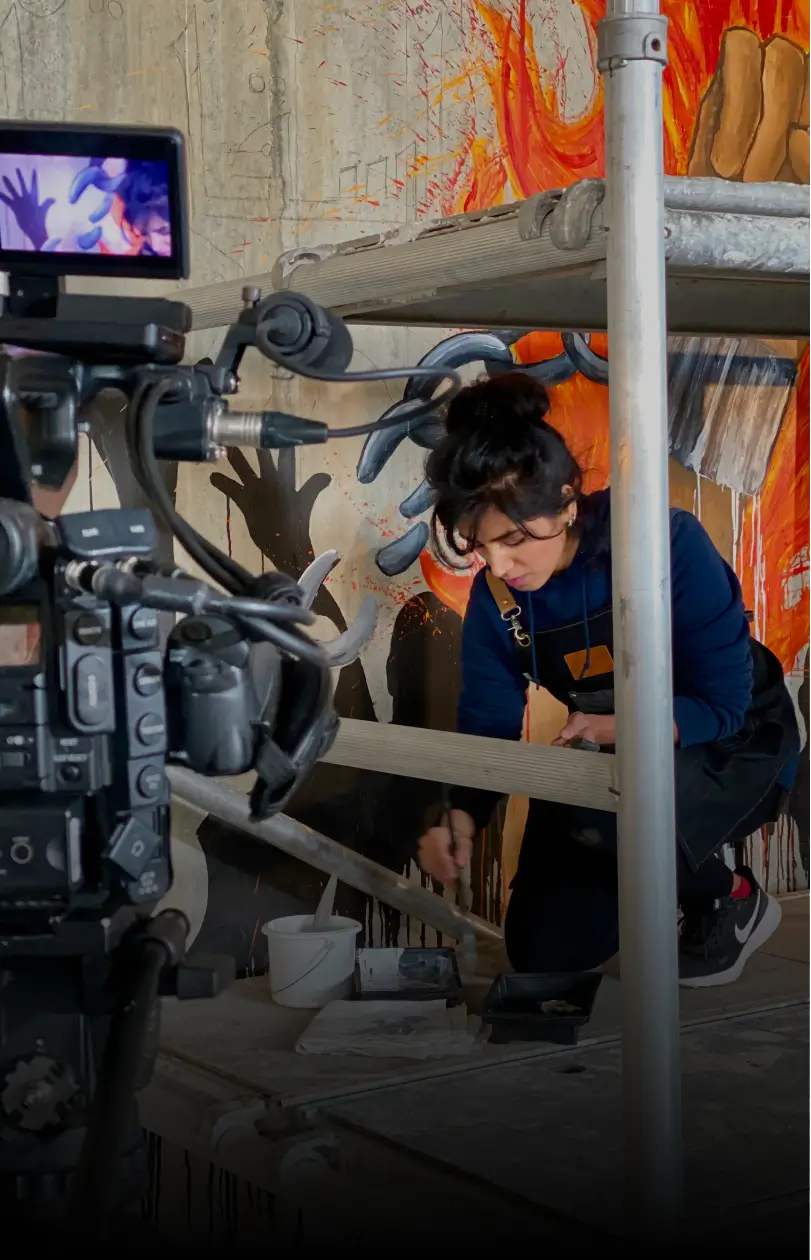 A woman artist painting a colorful mural on a wall while being filmed by a professional video camera.