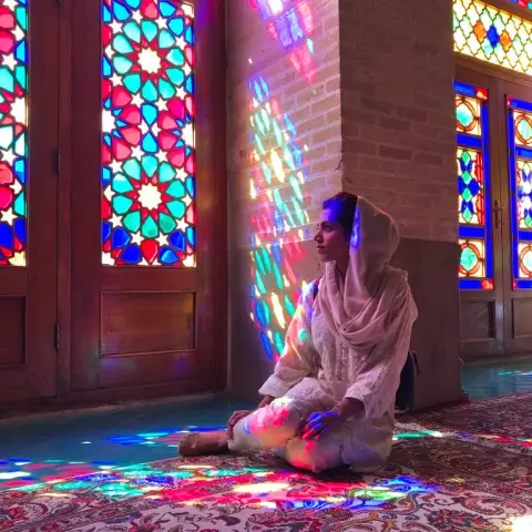 Woman in light pink traditional attire sitting on a patterned carpet with colorful stained glass light reflections on her and the walls.
