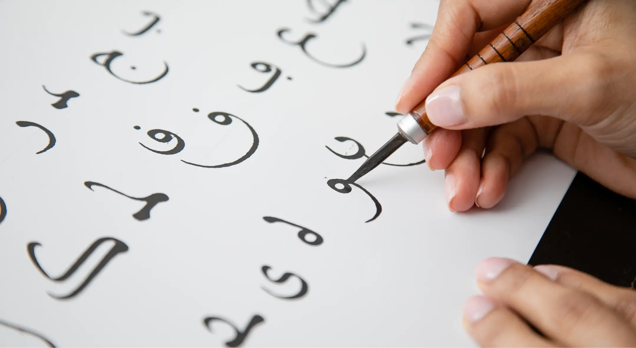Close-up of a hand using a calligraphy pen to write Arabic script on white paper.