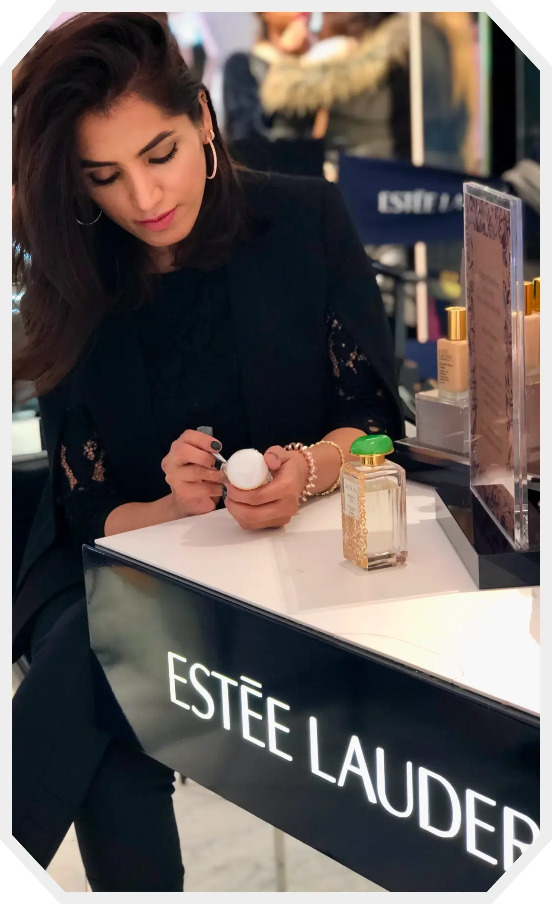 Woman writing an Arabic calligraphy script on a skincare product at an Estée Lauder counter featuring a perfume bottle with a green cap.
