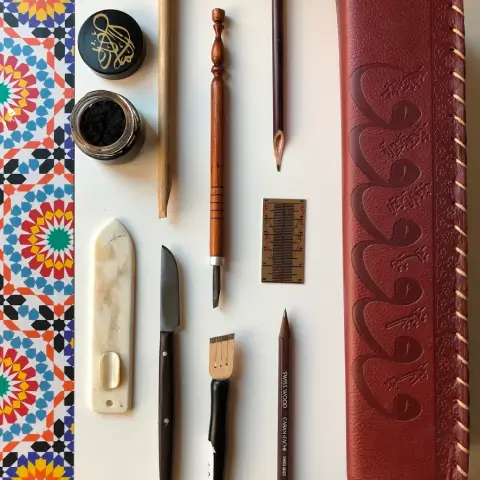 Set of calligraphy tools including pens, ink pot, knife, ruler, and leather case on a white surface with colorful geometric patterned border.