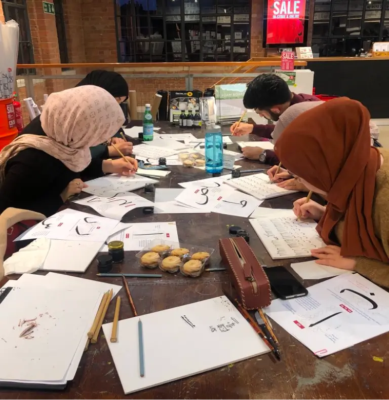 Four people wearing headscarves practicing Arabic calligraphy at a wooden table scattered with papers, ink pots, brushes, and snacks.