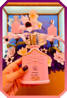 Hand holding a pink Creed Spring Flower perfume bottle with a background featuring pink flowers and two matching perfume bottles.