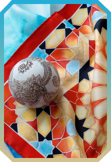 White ornament ball with intricate brown floral patterns resting on a colorful geometric patterned fabric with red, yellow, blue, and black shapes.